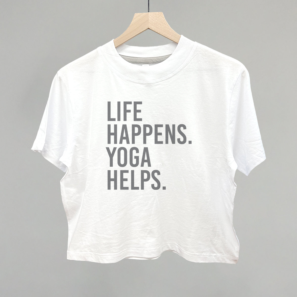 White short-sleeve shirt on a hanger with visible printed text, Life Happens. Yoga Helps.