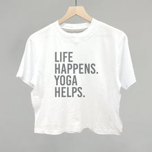 White short-sleeve shirt on a hanger with visible printed text, Life Happens. Yoga Helps.