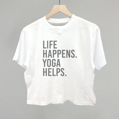 White short-sleeve shirt on a hanger with visible printed text, Life Happens. Yoga Helps.