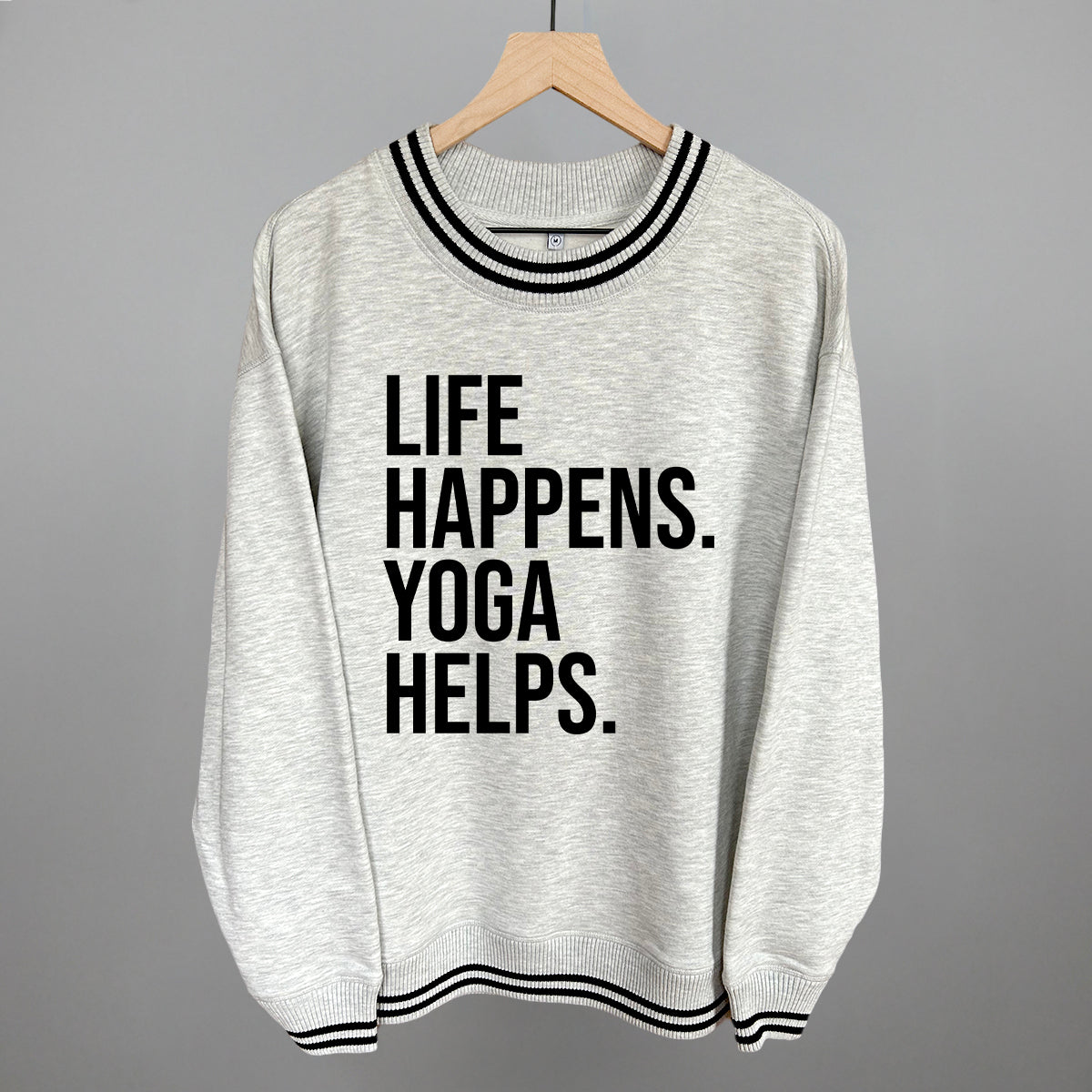 Light gray striped-trim sweatshirt on a hanger with visible printed text, Life Happens. Yoga Helps.