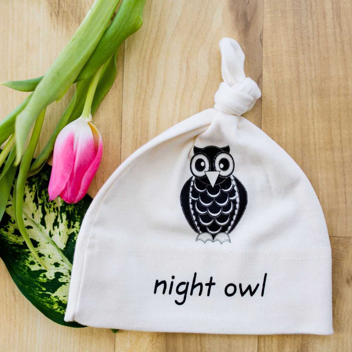 White knotted baby hat with black owl graphic and “night owl” text on a wood surface.