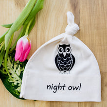 White knotted baby hat with black owl graphic and “night owl” text on a wood surface.
