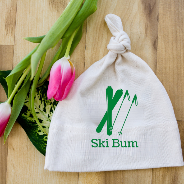 White knotted baby hat with green skier and mountain graphic and “Ski Bum” text beside pink tulips on a wood surface.
