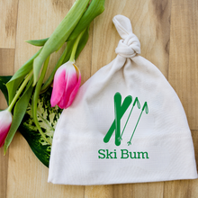 White knotted baby hat with green skier and mountain graphic and “Ski Bum” text beside pink tulips on a wood surface.
