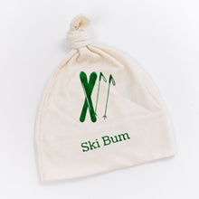White knotted baby hat with green skier and mountain graphic and “Ski Bum” text on a white background.