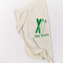 Natural cotton baby blanket with green skis and Ski Bum text folded on a white background