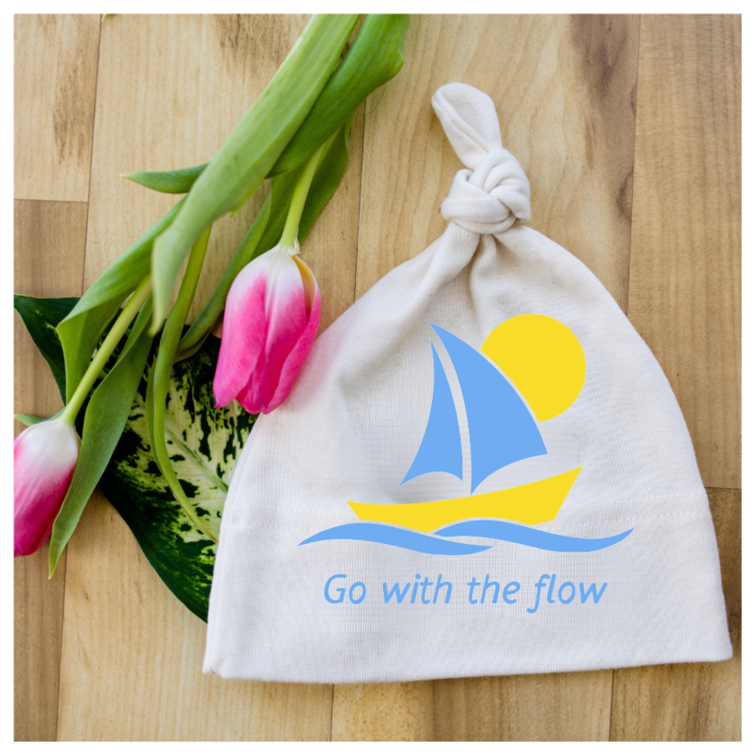 White knotted baby hat with blue sailboat graphic and “Go with the flow” text beside pink tulips on a wood surface.
