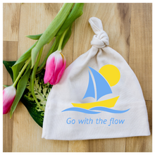 White knotted baby hat with blue sailboat graphic and “Go with the flow” text beside pink tulips on a wood surface.