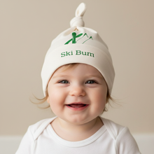 Baby wearing a white knotted hat with green skier and mountain graphic and “Ski Bum” text.
