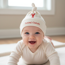 Baby wearing a white knotted hat with red lobster graphic and “Local Catch” text.