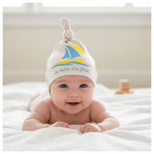 Baby wearing a white knotted hat with blue sailboat graphic and “Go with the flow” text.