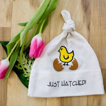 White knotted baby hat with yellow chick and “JUST HATCHED” text beside pink tulips on a wood surface.