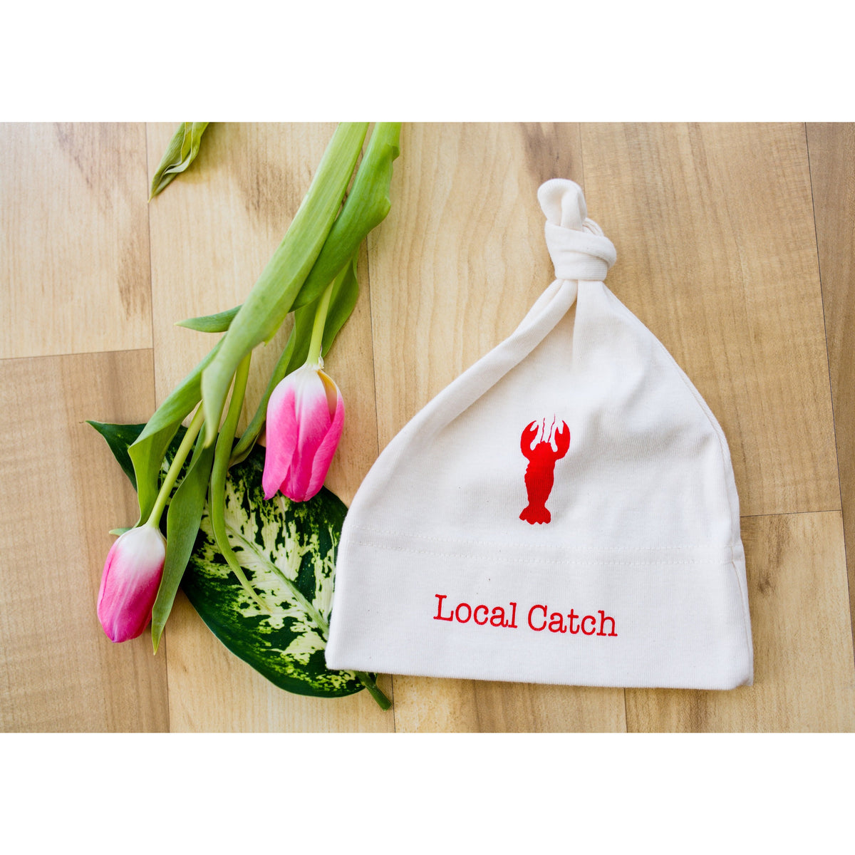 White knotted baby hat with red lobster graphic and “Local Catch” text beside pink tulips on a wood surface.