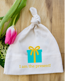Natural cotton knotted baby hat with a turquoise present and I Am the Present text beside pink tulips