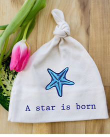 Natural cotton knotted baby hat with a blue starfish and A Star Is Born text beside pink tulips
