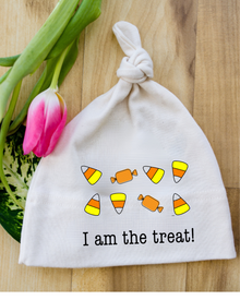 Natural cotton knotted baby hat with an I Am the Treat candy design beside pink tulips